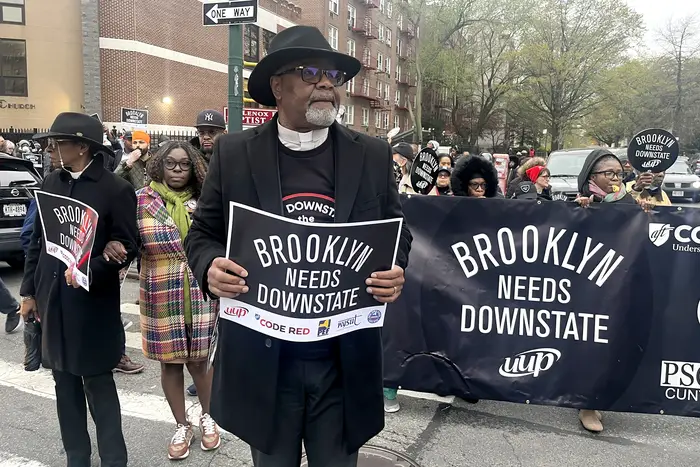 A man stands in front of a crowd, where he and several other people hold signs reading "Brooklyn needs Downstate"
